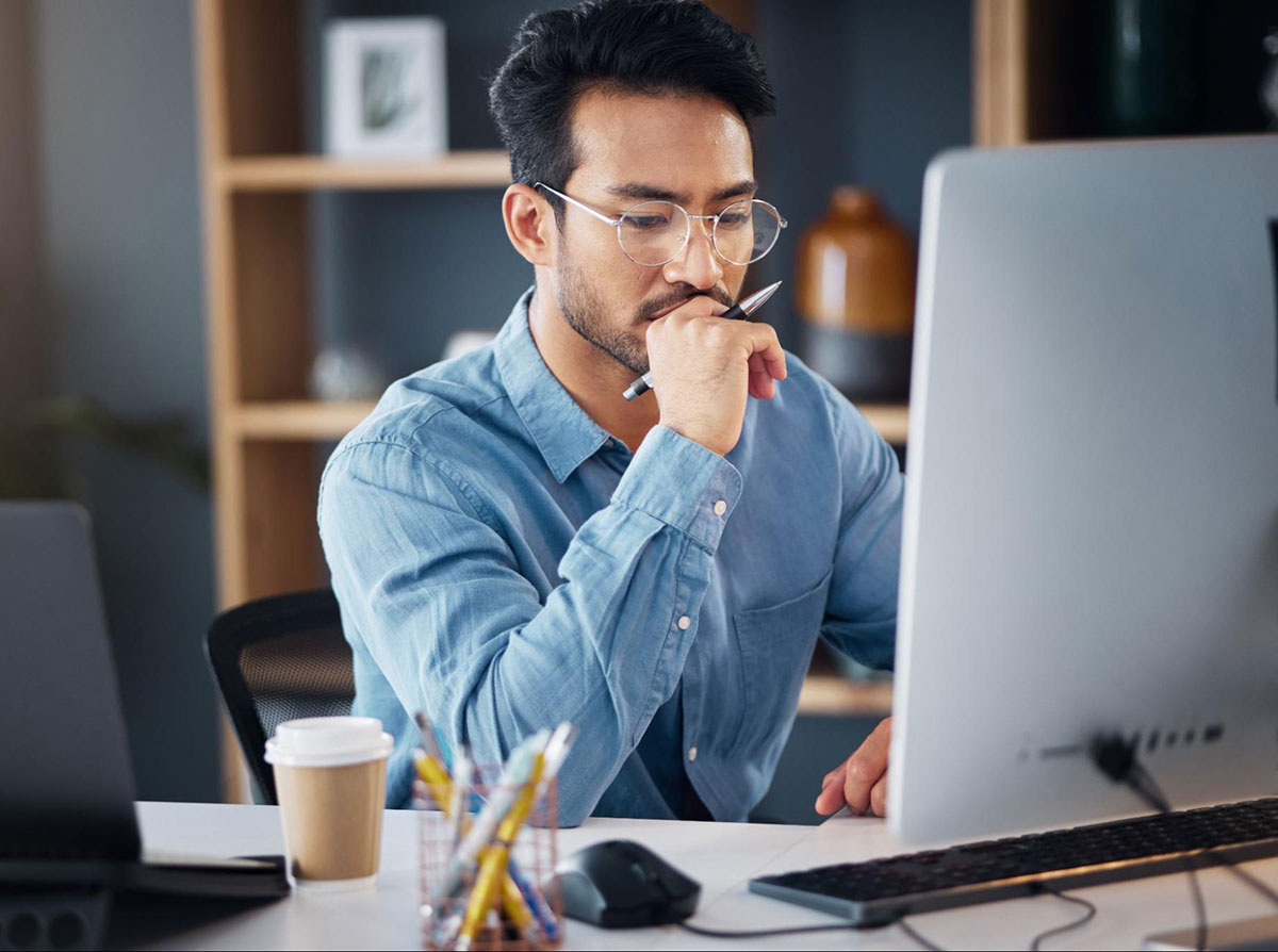 Person sitting at a desk with a desktop and laptop next to them. Person is thinking.