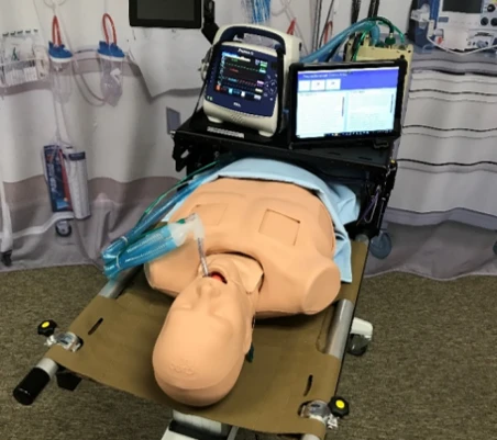 A medical training mannequin lying on a stretcher, intubated with airway tubing and connected to a portable patient monitor and an adjacent tablet displaying simulation data.