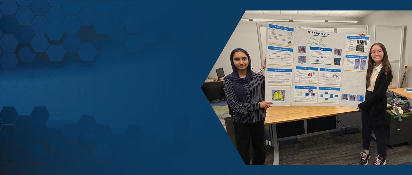 Two young women standing on either side of a tri-fold science presentation board about Kitware and Python scripting, set alongside a dark blue geometric hexagon background.