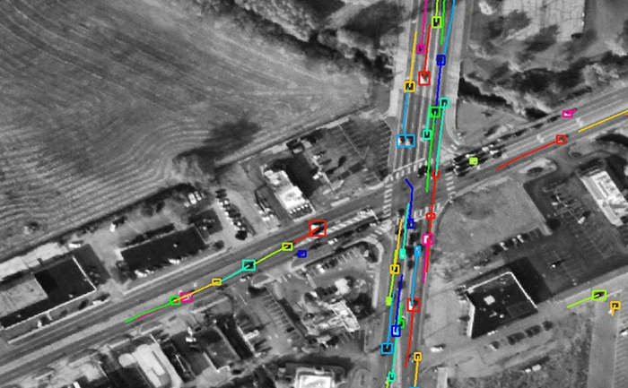 Aerial grayscale view of a road intersection with computer vision tracking boxes and colored trajectory lines following moving vehicles.