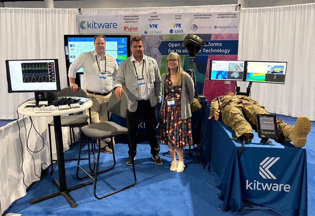 Three Kitware team members standing at an exhibition booth featuring medical simulation hardware, live Pulse Physiology Engine monitor displays, dual-screen software demos, and a trauma mannequin in military fatigue gear.